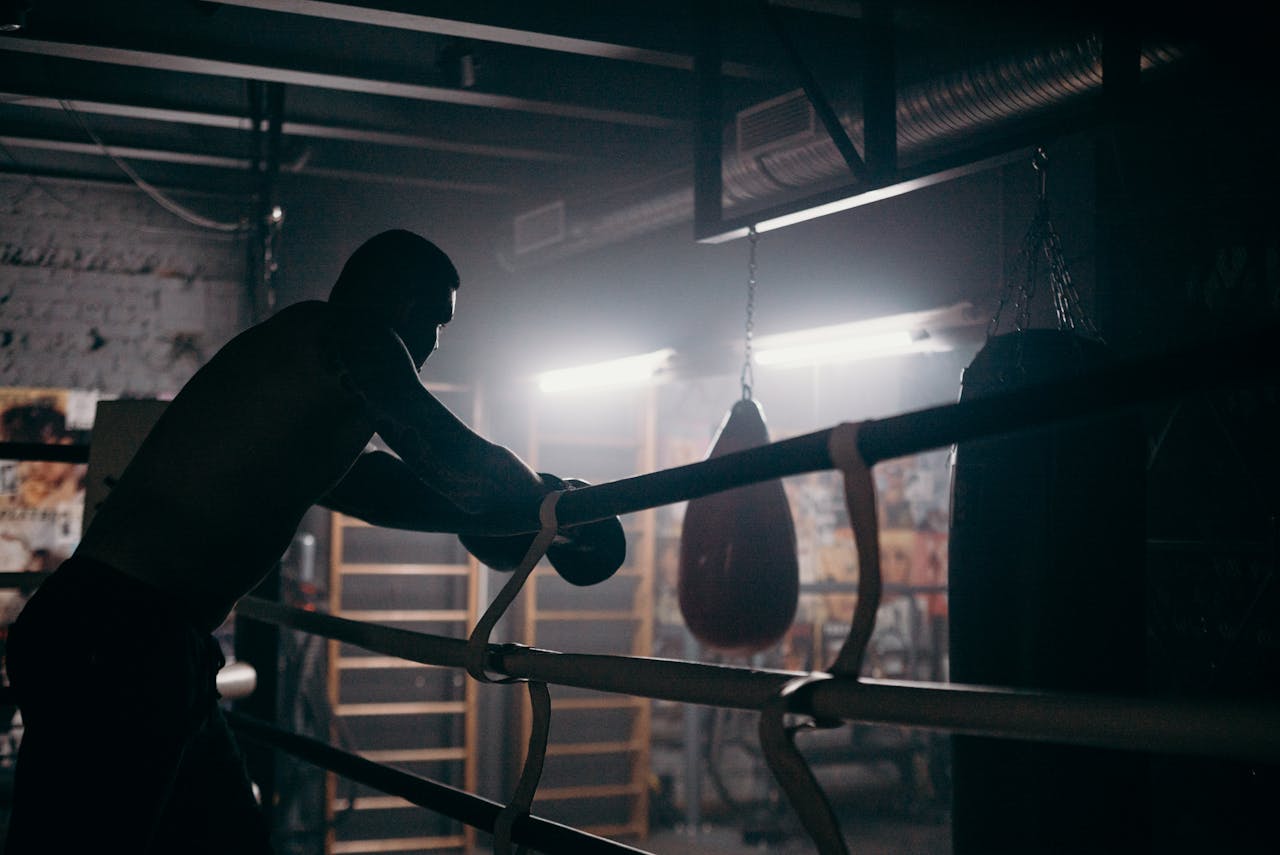 Moody silhouette of a boxer leaning in a dimly lit gym with punching bag and boxing ring.