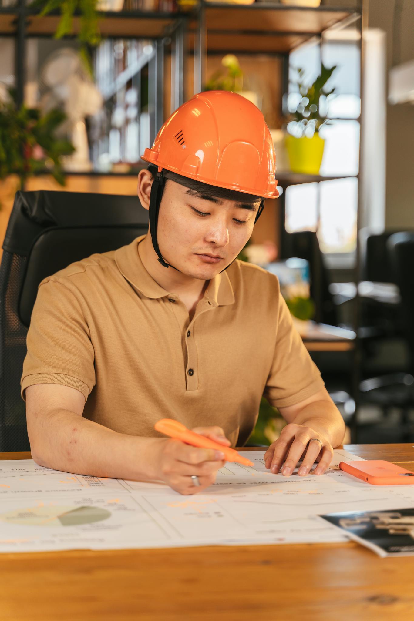 Asian engineer in hard hat examining blueprints in a modern office setting, focused and working.