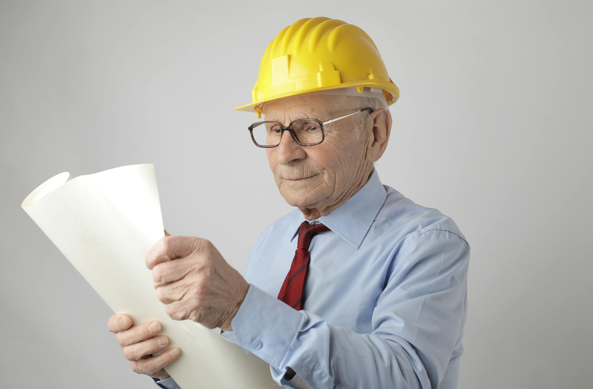 Gallery Elderly male engineer in hard hat reviewing blueprints in studio setting.
