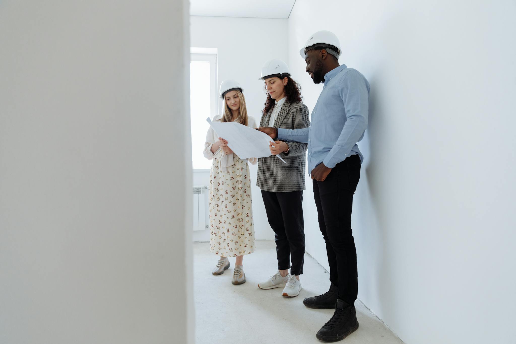 Gallery Three architects in hard hats review blueprints in a bright, empty white room.
